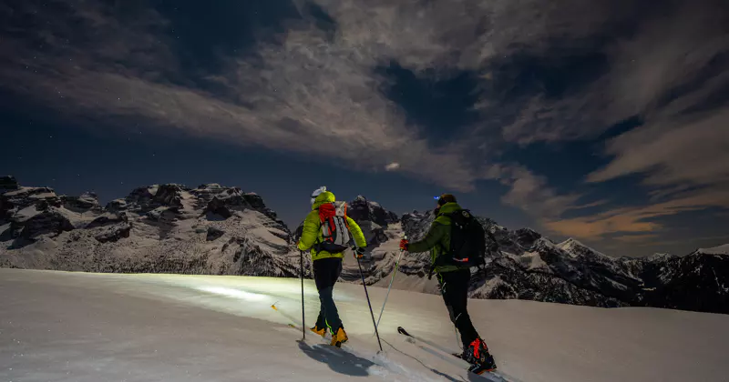 Twee mensen skiën ’s nachts op een besneeuwde berg omhoog, met hoofdlampen op, tegen een achtergrond van dramatische wolken en ruige bergtoppen.